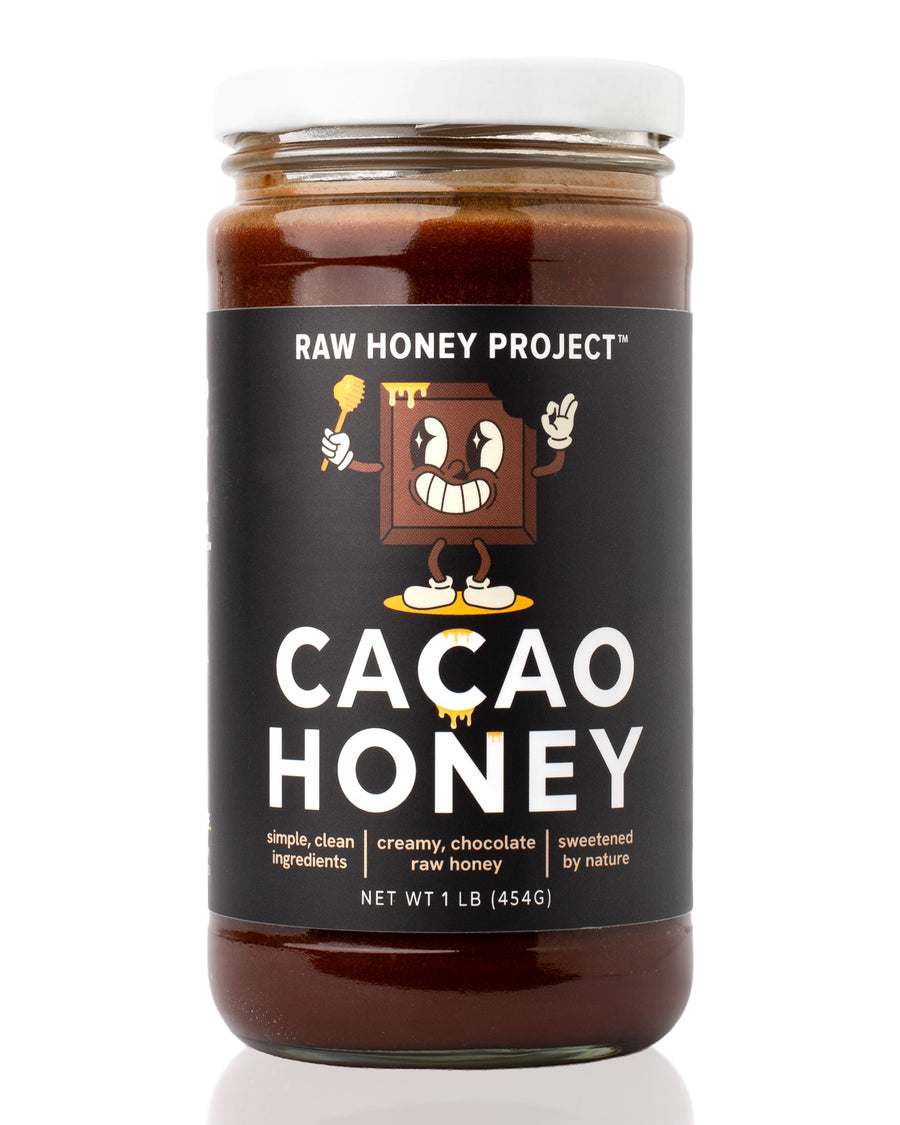 Glass jar of Raw Honey Project Cacao Honey, 1 lb, with a smiling cartoon chocolate bar mascot holding a honey dipper on a black label. Creamy dark chocolate-brown raw honey visible inside.