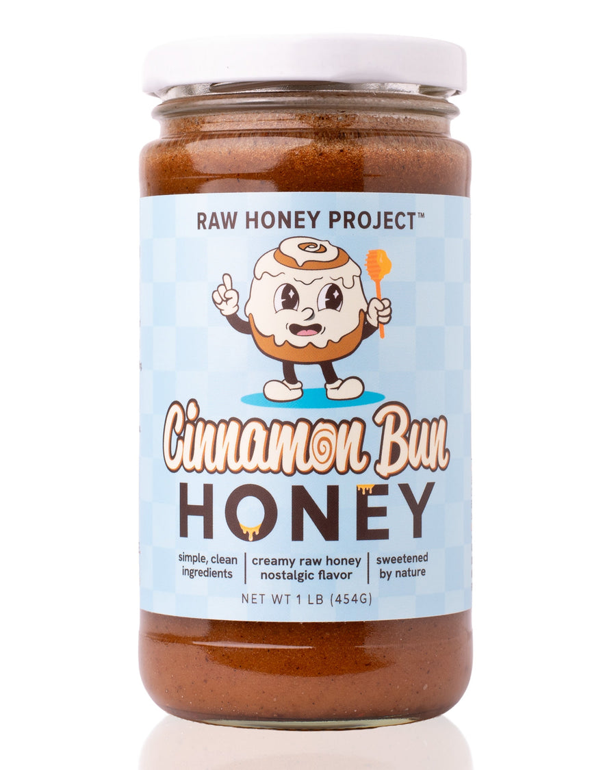 Glass jar of Raw Honey Project Cinnamon Bun Honey, 1 lb, with a smiling cartoon cinnamon roll mascot holding a honey dipper on a light blue checkered background. Creamy brown raw honey visible inside.