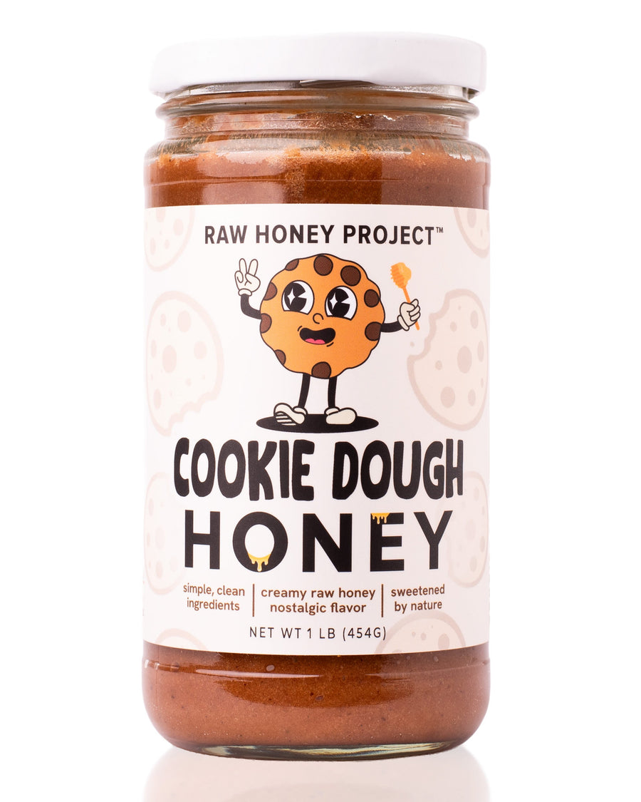 Glass jar of Raw Honey Project Cookie Dough Honey, 1 lb, with a smiling cartoon chocolate chip cookie mascot holding a honey dipper. Creamy brown raw honey visible inside.