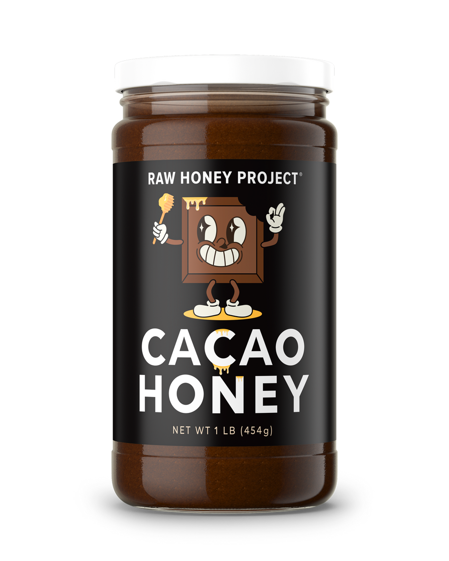 Glass jar of Raw Honey Project Cacao Honey, 1 lb, with a smiling cartoon chocolate bar mascot holding a honey dipper on a black label. Creamy dark chocolate-brown raw honey visible inside.