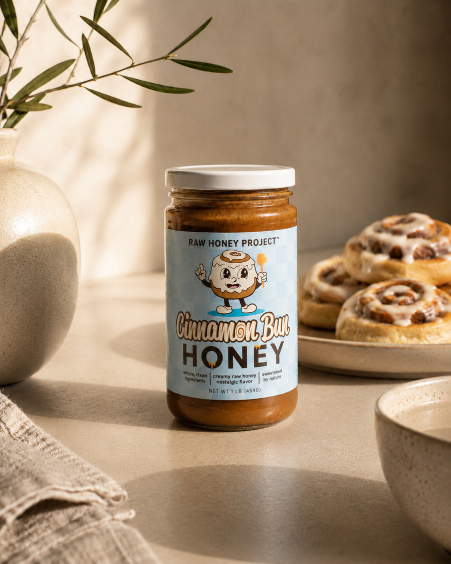 Jar of Cinnamon Bun Honey from Raw Honey Project on a table with cinnamon rolls in the background.