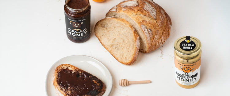 Overhead scene with artisan bread, a slice of toast topped with melted cacao honey, and jars of Raw Honey Project Cacao Honey and Cookie Dough Honey displayed on a light-colored kitchen surface.