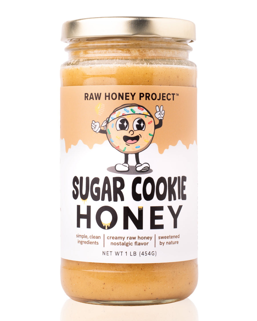 Glass jar of Raw Honey Project Sugar Cookie Honey, 1 lb, with a smiling cartoon sugar cookie mascot covered in sprinkles, holding a honey dipper on a tan and white label. Creamy pale-gold raw honey visible inside.